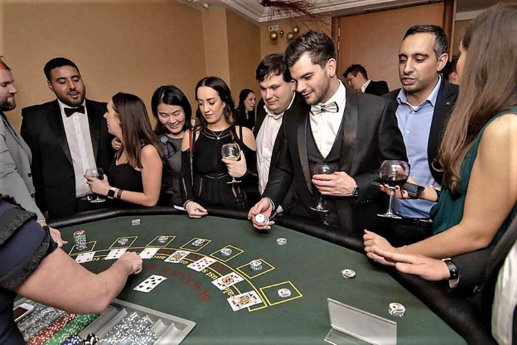 A group of elegantly dressed people in formal wear and black tie standing around a blackjack table, watching a man with a patterned bow tie roll dice on the green felt, capturing the excitement of a high-class Casino Royale.
