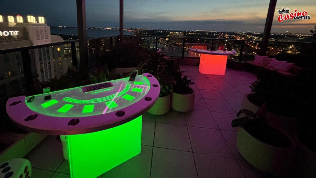 Illuminated green and pink LED blackjack tables set up on an outdoor rooftop terrace overlooking a city skyline at sunset for a glow-themed casino night.