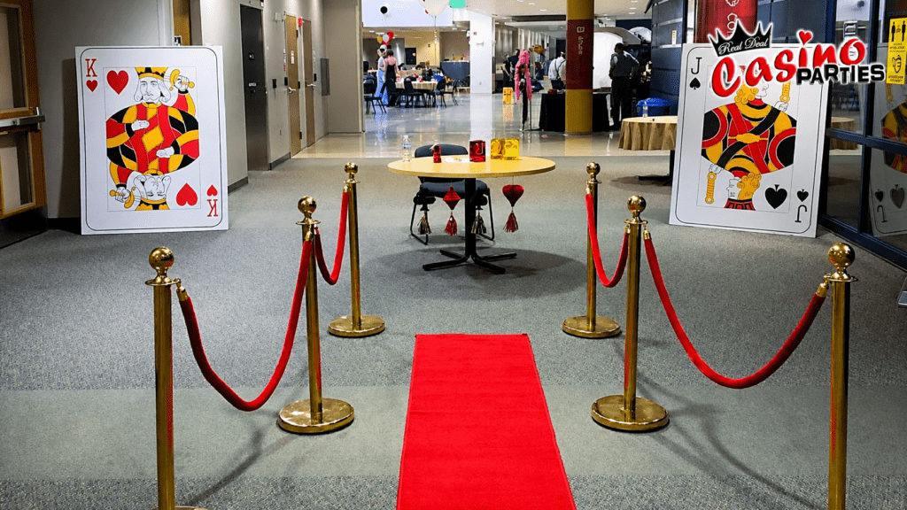 A red carpet entrance with gold stanchions and velvet ropes flanked by giant King of Hearts and Jack of Spades playing cards for a Hollywood-themed casino party.