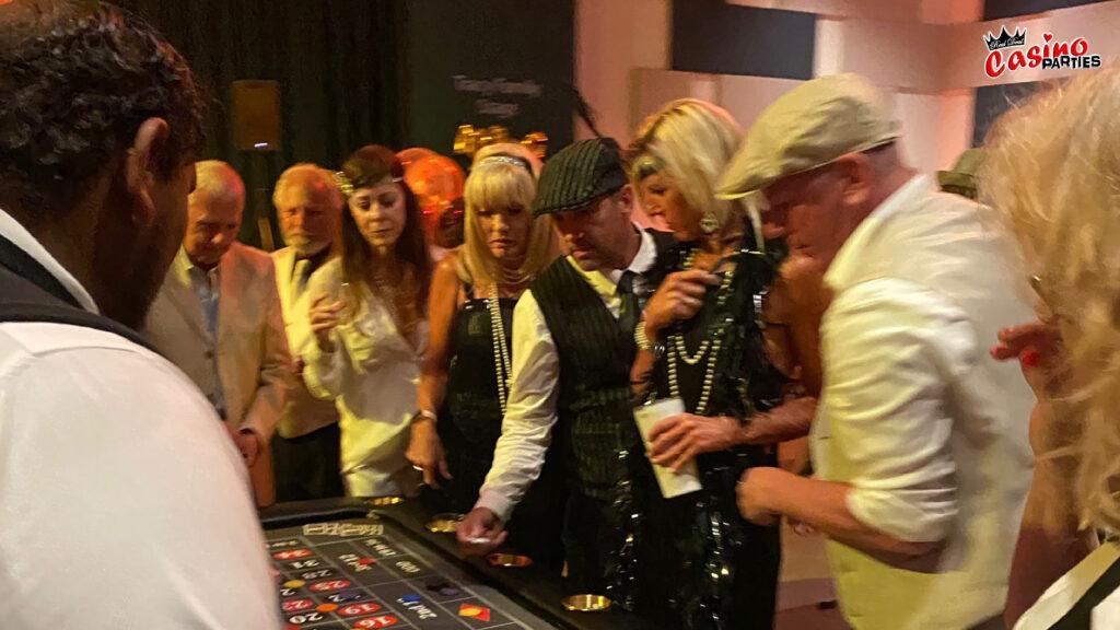 Guests in 1920s-style flat caps and flapper dresses gathered around a professional roulette table during a Prohibition Era themed casino party.
