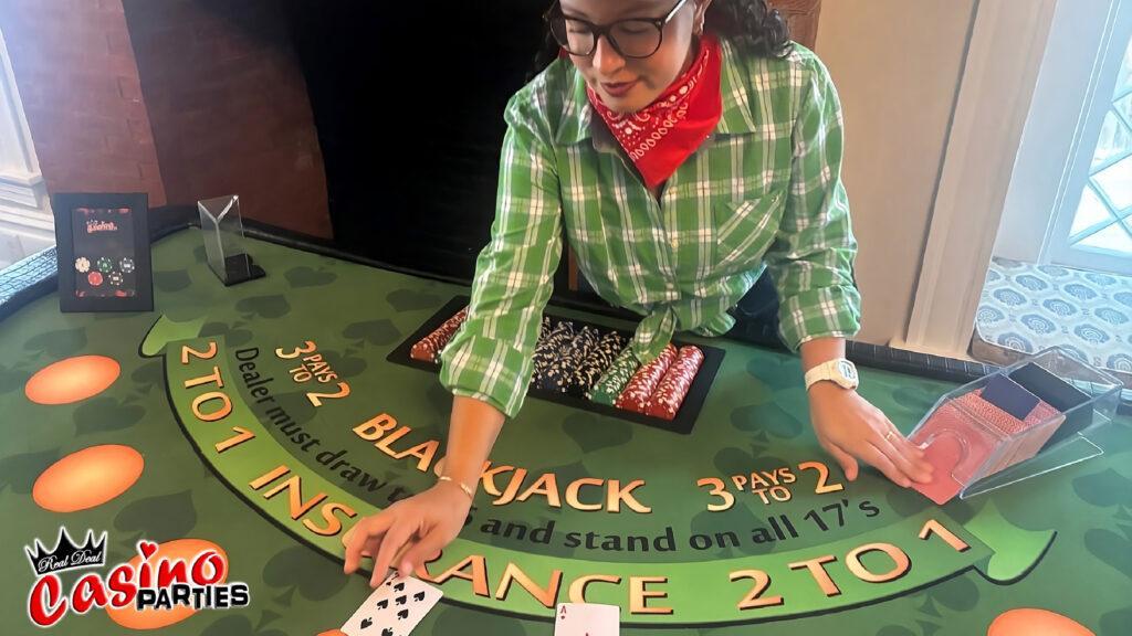 A female dealer in a green plaid shirt and red bandana dealing cards at a professional blackjack table for a Wild West casino night event.