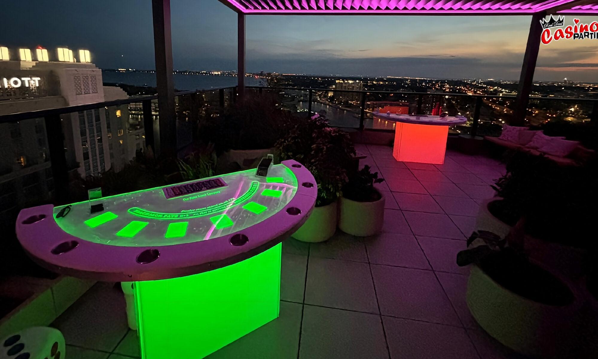 Vibrant green and red LED blackjack and casino tables on a rooftop terrace overlooking a city skyline at sunset.