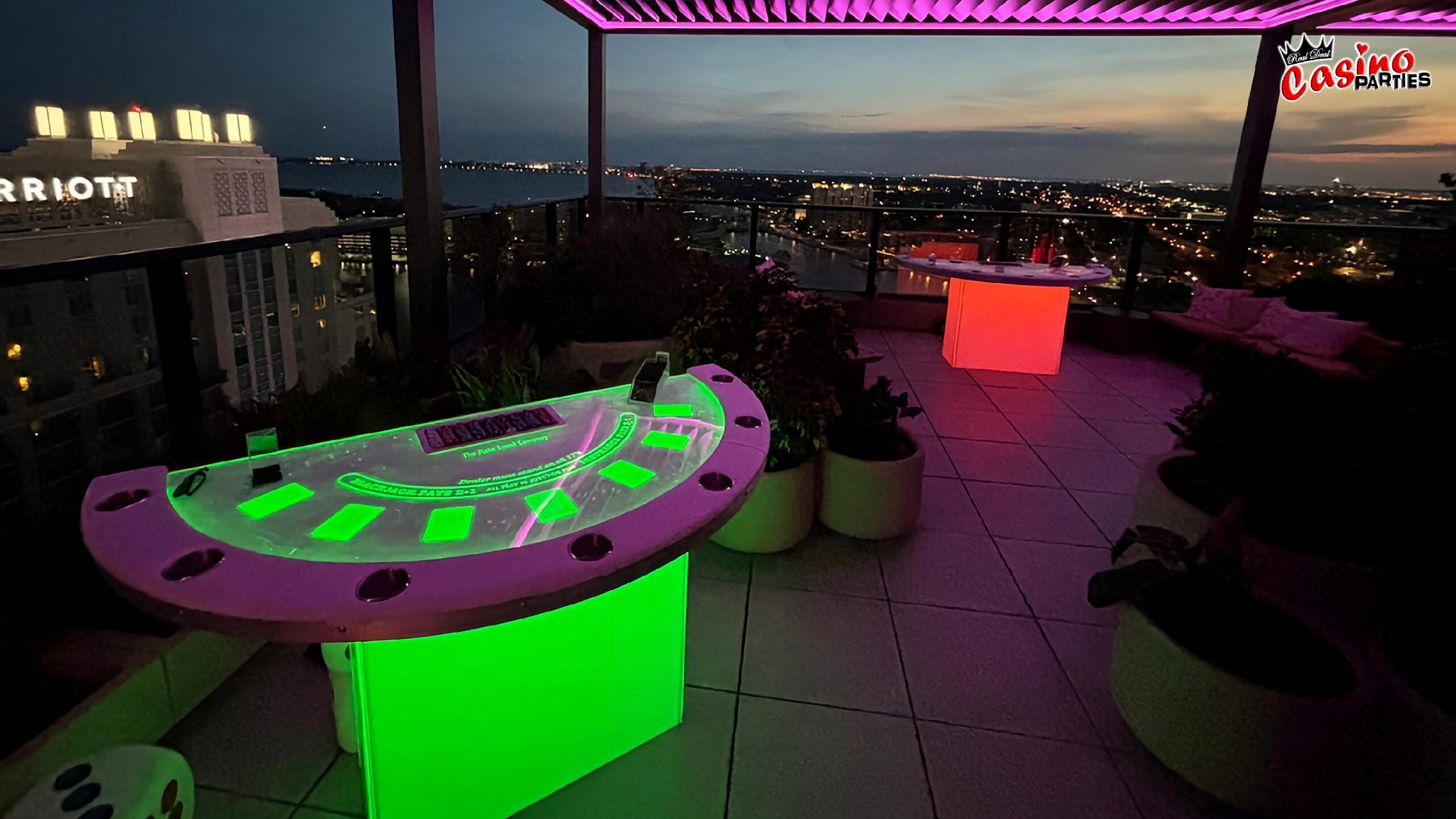 Vibrant green and red LED blackjack and casino tables on a rooftop terrace overlooking a city skyline at sunset.